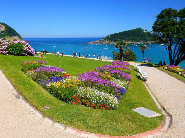 Ein Park mit bunten Blumen, grünem Gras, Bäumen, Bänken, Menschen, die spazieren gehen, Booten auf dem Wasser, Gebäuden und einem klaren blauen Himmel im Hintergrund.