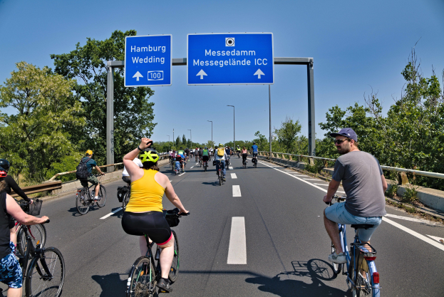 Gruppe von Radfahrern mit Helmen auf einer Straße mit Geländer und Bäumen auf beiden Seiten, Laternen im Hintergrund und einem klaren blauen Himmel, mit einem Schild, das eine Radtour in Hamburg anzeigt.