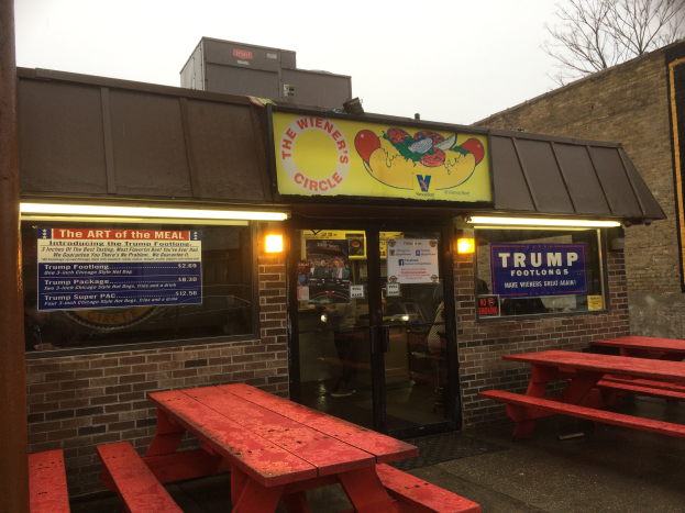 Außensitzbereich eines Restaurants mit roten Picknicktischen, Glaswänden und Postern an den Innenwänden, vor Buildings, Bäumen und einem klaren blauen Himmel mit wenigen Menschen drinnen.