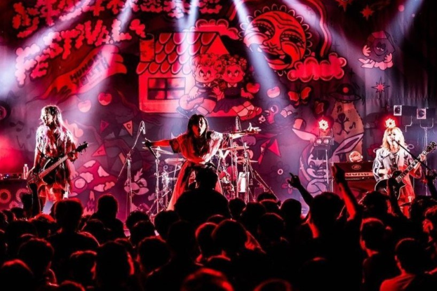 Eine Live-Aufführung der japanischen Rockband Korn auf der Bühne, mit Bandmitgliedern, die Gitarren spielen und in Mikrofone singen, einer beleuchteten Wandmalerei im Hintergrund und einem sichtbaren Publikum unten.