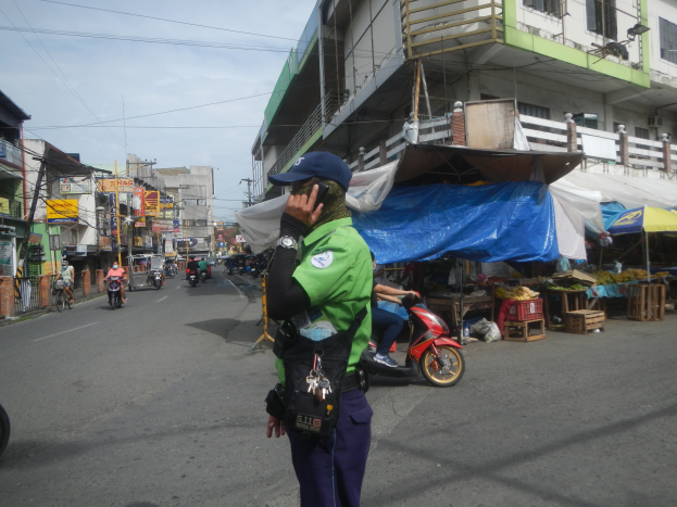 Ein Polizist in grüner und blauer Uniform und blauer Mütze steht an der Straße, spricht in ein Handy, mit Fahrzeugen im Hintergrund, Ständen mit Gegenständen rechts und einer bewölkten Himmel.