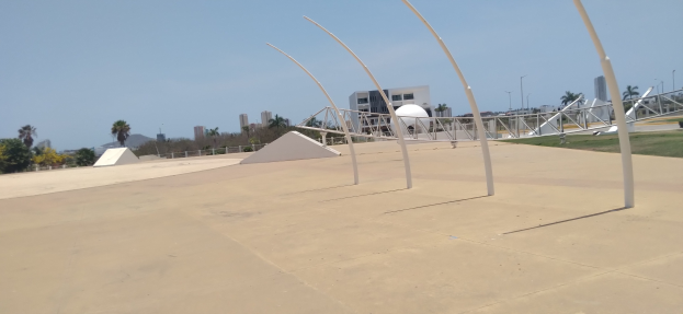 Eine Skateranlage mit einem zentralen Metallbau, umgeben von Pfählen, Gras, einem Zaun, Bäumen, Gebäuden, Straßenlaternen und einem bewölkten Himmel.