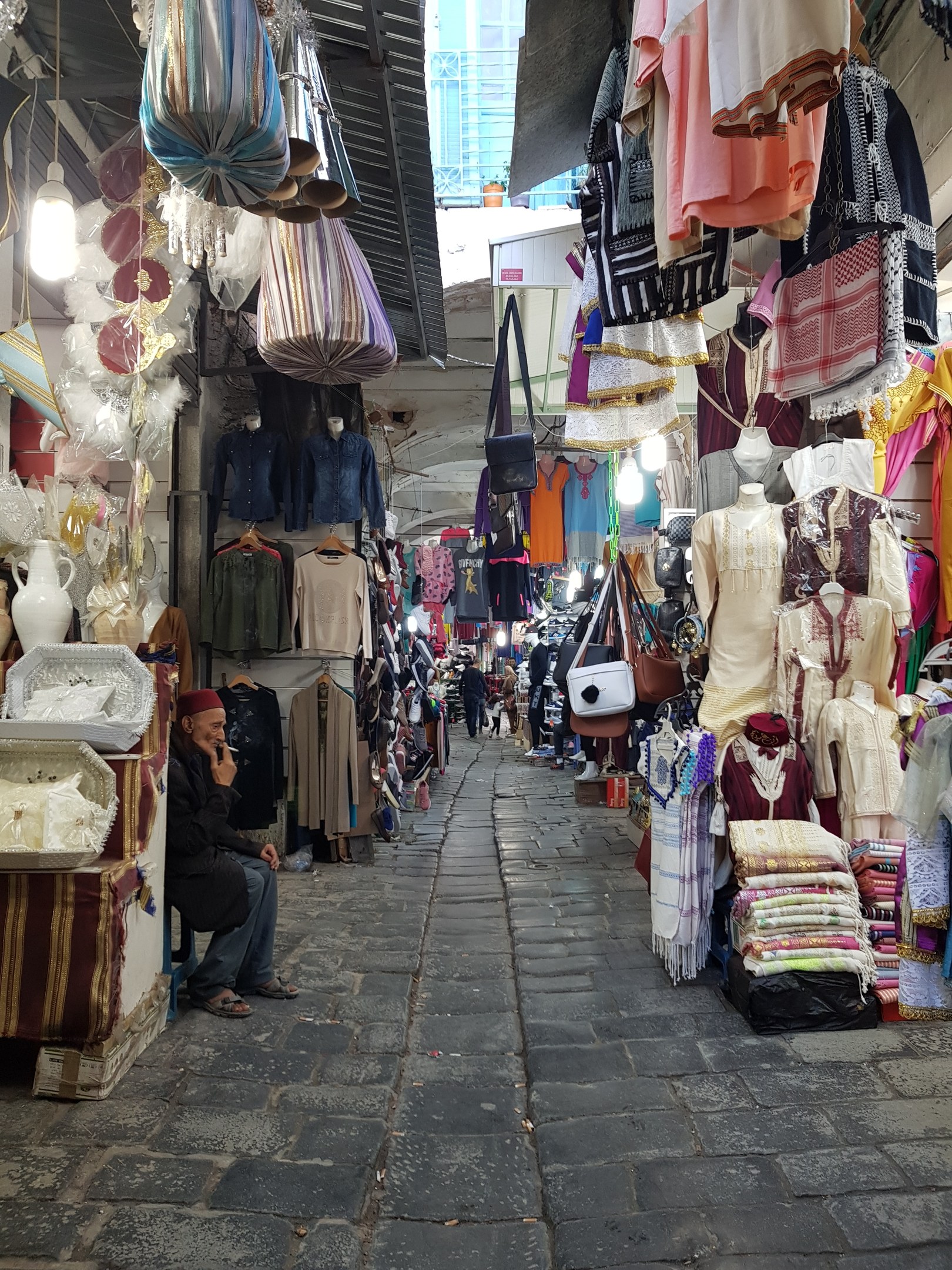 Ein belebter Straßenmarkt in der Altstadt von Jerusalem mit Ständen, die Kleidung, Taschen, Lampen und andere Artikel verkaufen, während Menschen umhergehen und stöbern, mit einem Gebäude im Hintergrund.