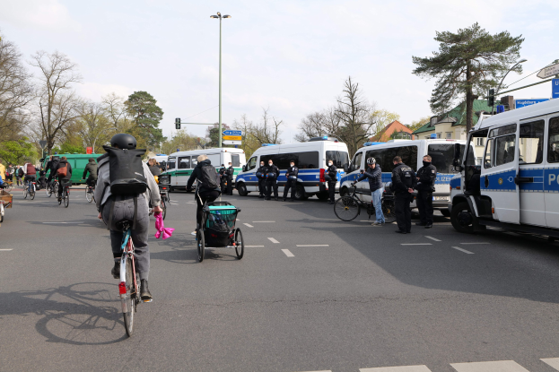 Eine Gruppe von Menschen, die auf Fahrrädern eine Straße entlangfahren, mit Polizeiwagen und anderen Fahrzeugen, einige tragen Helme und haben Taschen oder schieben Kinderwagen, vor einer Kulisse aus Bäumen, Gebäuden und Verkehrsinfrastruktur.