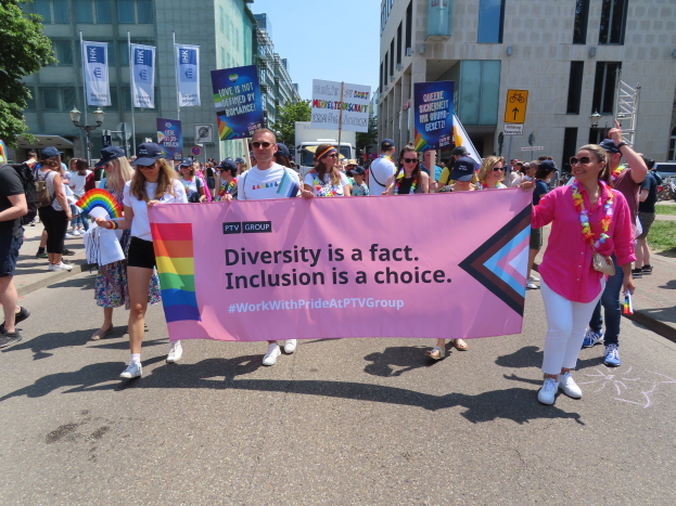 Eine Gruppe von Menschen marschiert auf einer Straße, hält ein Banner mit der Aufschrift "Diversität ist eine Tatsache, Inklusion ist eine Wahl" in den Händen, einige tragen Mützen und Schutzbrillen und andere halten zusätzliche Banner, vor einem Hintergrund aus Gebäuden, Bäumen und einem klaren Himmel.