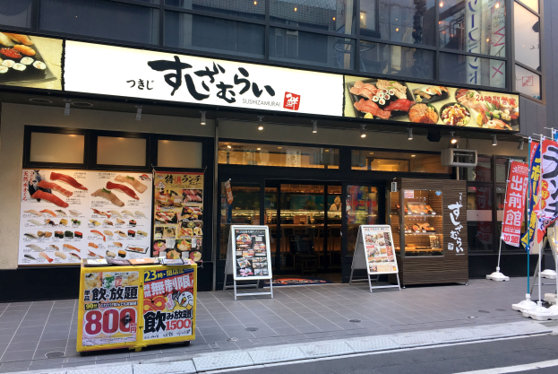 Außenansicht eines Sushi-Restaurants in Tokyo mit Glaswänden, Essensauslagen, Schildern und einer Straße im Vordergrund.