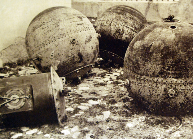 Drei große Metallbomben auf dem Boden vor einer Wand mit einem Fenster im Hintergrund, dargestellt in Schwarz-Weiß.