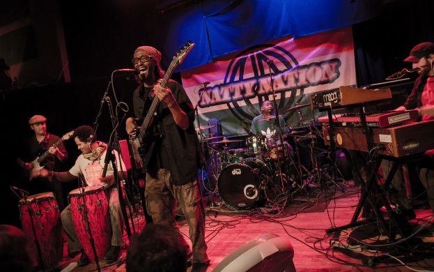 Eine Live-Band performt auf der Bühne mit Mikrofonen und Ständern, während das Publikum unten zuschaut; ein Banner ist im Hintergrund zu sehen.