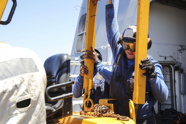 Zwei Männer in blauen Uniformen und Schutzausrüstung arbeiten an einem gelben Gabelstapler, einer hält ein Metallobjekt, während im Hintergrund ein Fahrzeug und der Himmel zu sehen sind.