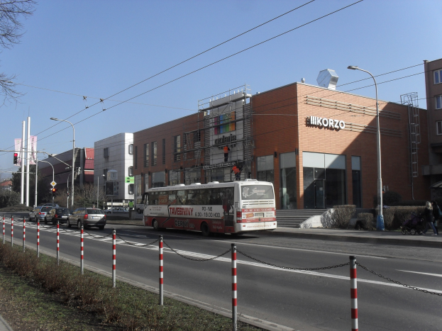 Ein Bus fährt auf einer Straße mit hohen Gebäuden, Strommästen und Straßenlaternen, mit Kraftfahrzeugen, Füülern und Vegetation unter einem klaren blauen Himmel.