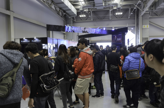 Eine Gruppe von Menschen steht vor einer Menge bei einer Gaming-Messe, mit einigen Besuchern, die Taschen, Schilder, Deckenlichter und Rohre im Hintergrund tragen.