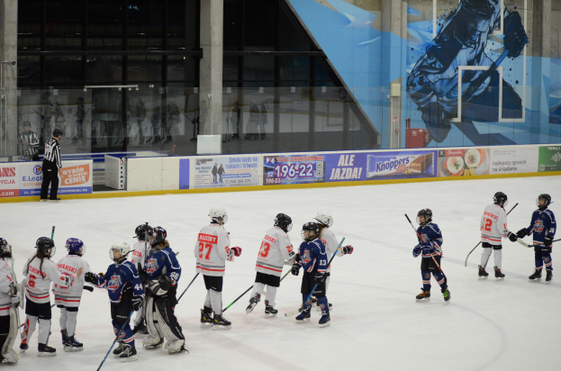 Gruppe junger Menschen beim Eishockey auf einer Indoor-Eisfläche, mit Helmen und Stöcken, mit einer Person auf der linken Seite und Bannern, Säulen und Glaswänden im Hintergrund.