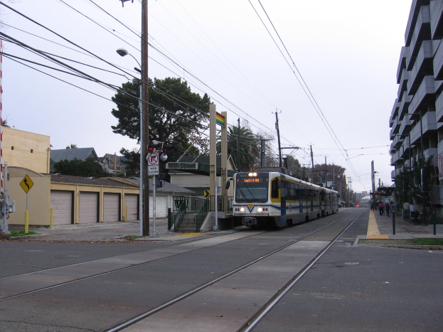 Straßenbahn auf einer Stadtstraße mit Hochhäusern, Strommasten, Fußgängern, Bäumen und einem klaren blauen Himmel.