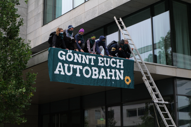 Gruppe von Menschen auf einem Gebäude mit einer Fahne, mit einem Baum links und Glasfenstern im Hintergrund, wahrscheinlich protestierend für die deutsche Regierung.
