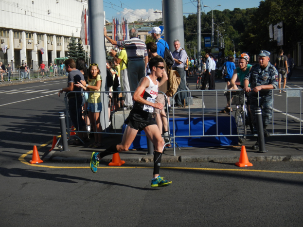 Ein Mann beim Laufen eines Marathon auf einer Stadtstraße, mit Zuschauern und Radfahrern auf dem Gehweg, unter einem bewölkten Himmel.