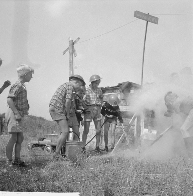 Schwarzes und weißes Bild von Kindern in Helmen, die sich um einen Hydranten in einem grasbewachsenen Feld versammelt haben, einige halten Gegenstände, mit Strommasten und einem Schild im Hintergrund.