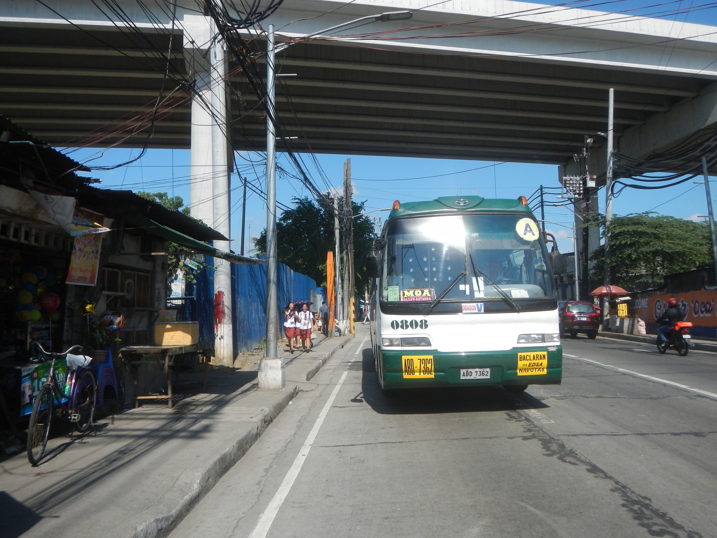 Ein grüner und weißer Bus fährt auf einer Straße mit Strommasten und -leitungen an Geschäften und Fußgängern vorbei, die eine Brücke überqueren, mit Fahrrädern, die an der Straße geparkt sind, und Bäumen im Hintergrund bei klarem Himmel.