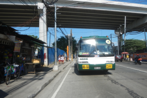 Ein grüner und weißer Bus fährt auf einer Straße mit Strommasten und -leitungen an Geschäften und Fußgängern vorbei, die eine Brücke überqueren, mit Fahrrädern, die an der Straße geparkt sind, und Bäumen im Hintergrund bei klarem Himmel.