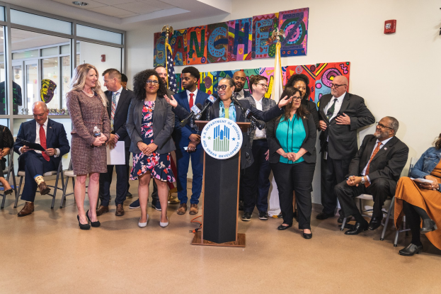 Gruppe von Menschen vor einem Rednerpult mit Mikrofon und einer Tafel mit dem Text "Sancho City Councilman Joins City Council", mit sitzenden und stehenden Teilnehmern, Flaggen, einem Feuerlöscher und Deckenlampen im Hintergrund.