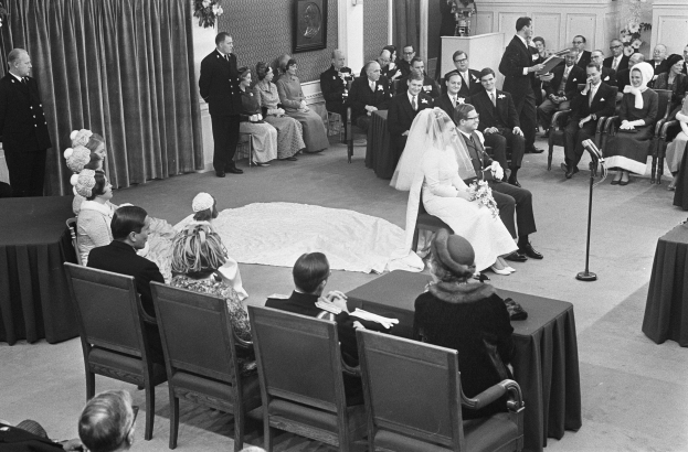 Schwarz-weiß-Foto einer Hochzeitszeremonie mit sitzenden und stehenden Menschen, einem Mann mit Mikrofon, einer Frau mit Blumenstrauß, einem Podium mit Mikrofon, Blumenschmuck, einem Vorhang, einem gerahmten Foto an der Wand und einer Tür im Hintergrund.