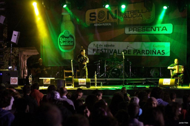 Publikum, das Künstler auf einer Bühne mit Musikinstrumenten, Lautsprechern, Mikrofonen, einem Hintergrund und Scheinwerfern beobachtet, mit einer Flasche und anderen Gegenständen im Hintergrund.