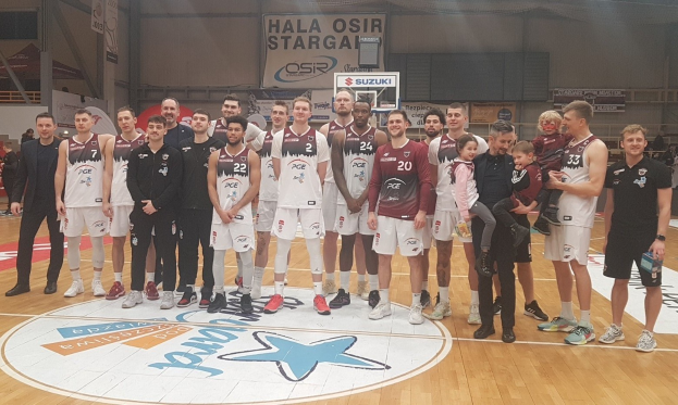 Mannergruppe mit Kindern auf einem Basketballfeld mit Schildern "Hala Osir Starga", sitzende Zuschauer, ein Basketballkorb, Metallstangen, Deckenbeleuchtung und eine Wand im Hintergrund.