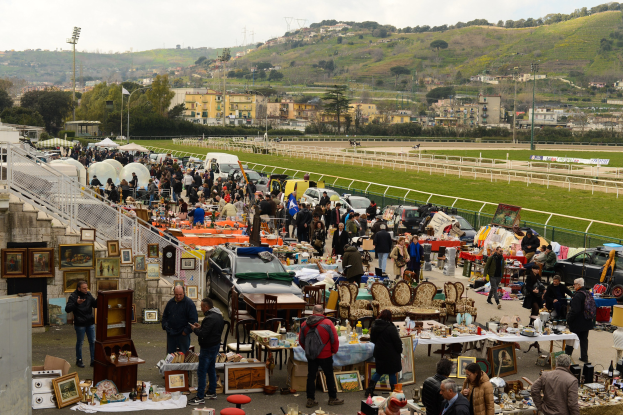 Große Gruppe von Menschen auf einem Flohmarkt mit Tischen, auf denen Gegenstände wie Foto Rahmen und Stühle ausgelegt sind, umgeben von parkenden Fahrzeugen, Geländern, Treppen, Bäumen, Gebäuden, Laternenmasten, Hügeln und einem bewölkten Himmel.