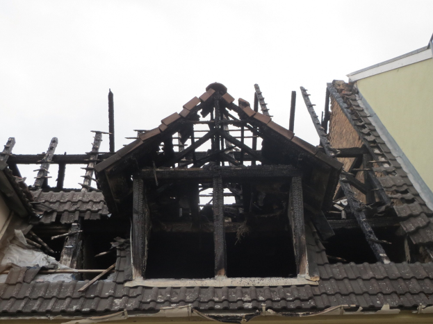 Brandgeschädigtes Gebäude mit verkohltem Dachschutt und verstreuten Holzstücken während der Abrissarbeiten, vor einem Himmel-Hintergrund.