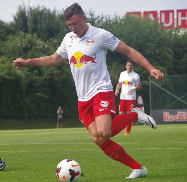 Ein professioneller Fußballspieler schießt einen Ball auf einem grünen Feld mit Bäumen und einem klaren blauen Himmel im Hintergrund, wobei "RB Leipzig" auf einem Schild zu sehen ist.