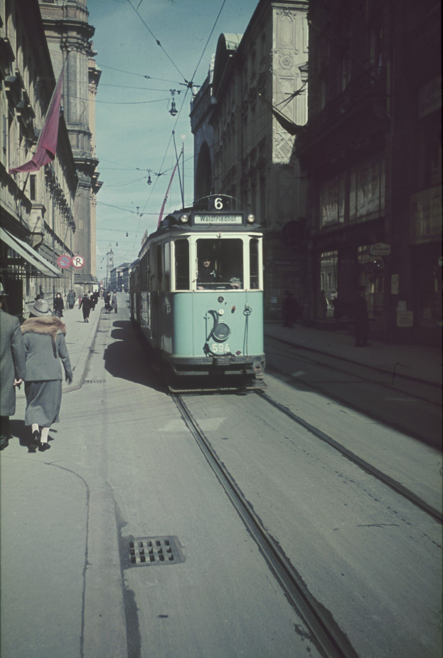 Altes Schwarz-Weiß-Foto einer Straßenbahn, die auf einer Stadtstraße mit Fußgängern auf dem linken Gehweg und Gebäuden, Strommasten, Drähten und einem klaren blauen Himmel im Hintergrund fährt.