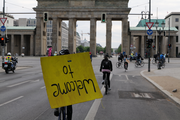 Eine Gruppe von Menschen, die auf Fahrrädern vor dem Brandenburger Tor in Berlin, Deutschland, die Straße entlangfahren, wobei eine Person ein gelbes Schild mit Text hält.