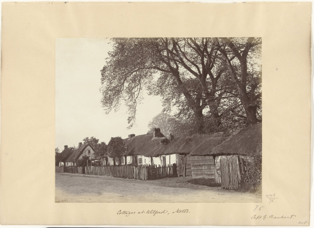 Schwarzes und weißes Foto eines Dorfs mit Reetdachhäusern, einer Straße, einem Zaun, Bäumen und einem Himmel im Hintergrund, mit Text unten.