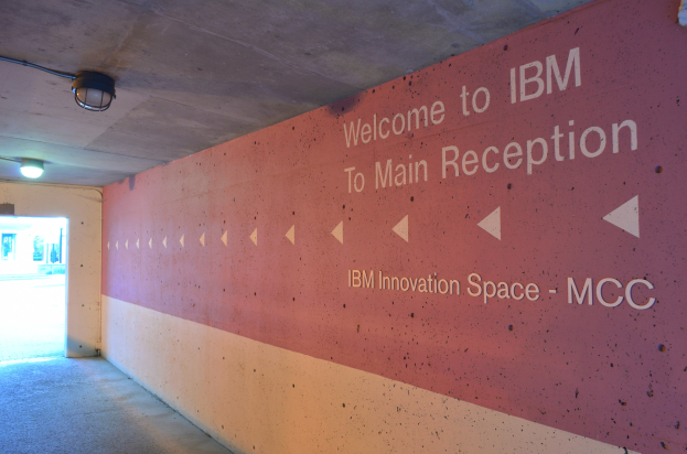 Eine Wand mit den Worten "Willkommen bei IBM" beleuchtet von Deckenlampen, mit einer Tür im Hintergrund beschriftet "IBM Innovation Space MCC."