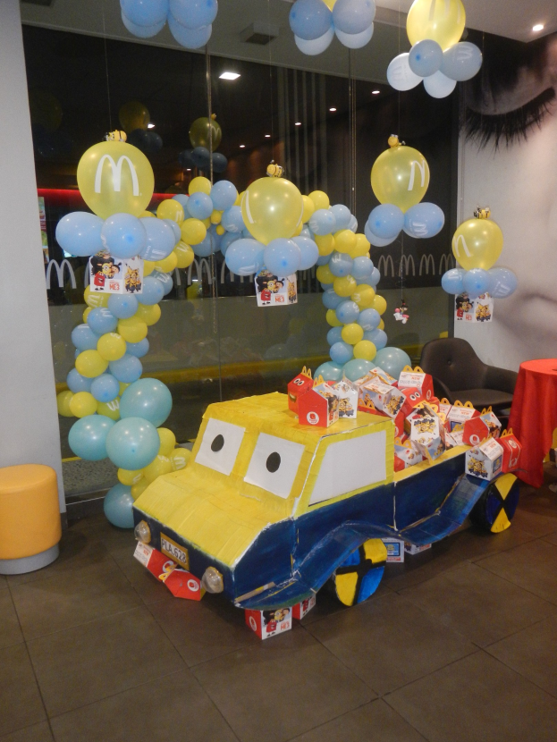 Eine McDonald's-Geburtstagsparty-Szene mit einem gelben und blauen Lastwagen, der mit Ballons und anderen Dekorationen geschmückt ist, einem rot gedeckten Tisch mit Gegenständen, Hockern, einer Glaswand und Deckenlampen, die eine festliche Atmosphäre schaffen.