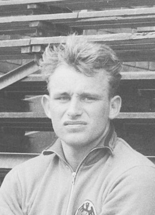 Schwarzes und weißes Foto eines Mannes in einer Jacke, der vor einem Haufen Holzplanken steht, der als legendärer deutscher Fußballspieler gilt, der im Alter von 89 Jahren verstorben ist.