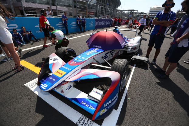Formula E Rennauto mit weißer, blauer, roter und schwarzer Lackierung auf einer Strecke geparkt, umgeben von Menschen, mit einem Schirm im Vordergrund und Rennstreckenabsperrungen, Gebäuden, Bäumen und einem klaren blauen Himmel im Hintergrund.