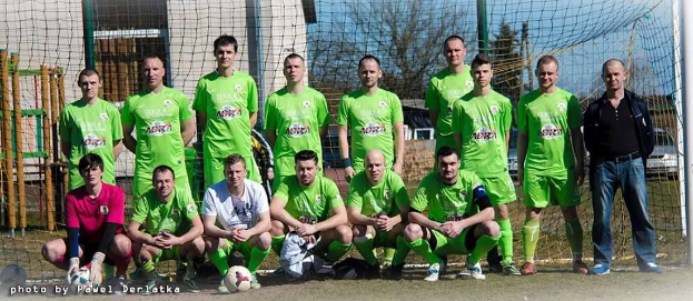 Eine Gruppe von Männern in grünen T-Shirts steht und hockt auf einem Fußballfeld mit einem Ball davor, ein Torpfosten rechts daneben und eine Hintergrundlandschaft aus Bäumen, Gebäuden, Pfählen, Fahrzeugen und Himmel.