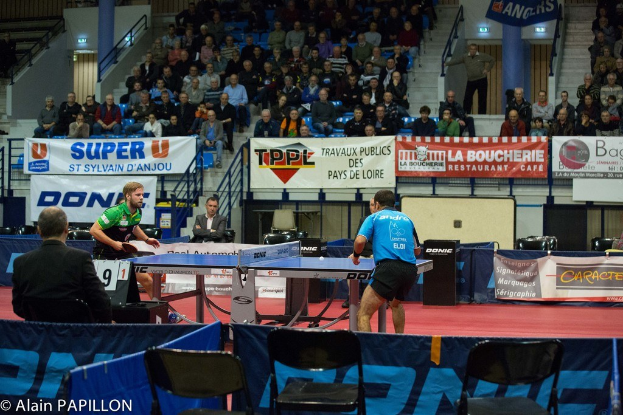 Indoor-Tischtennis-Match mit zwei Spielern, die gegeneinander antreten, sowie einigen stehenden Zuschauern, einer sitzenden Menge, leeren Stühlen und Werbeplakaten in einer Stadionatmosphäre.
