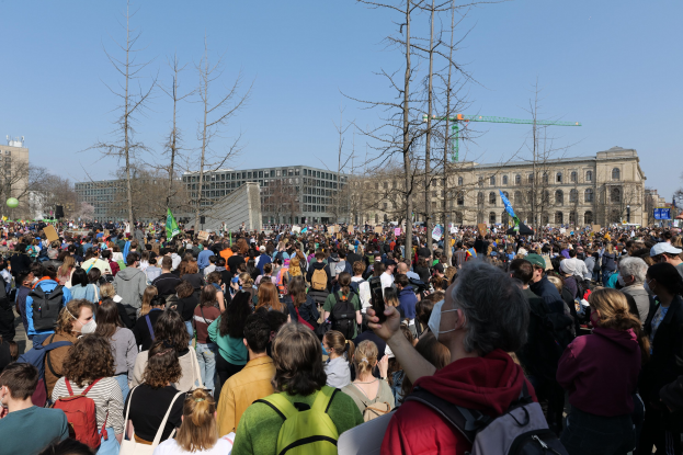Eine große Menschenmenge bei einer Klimaprotestveranstaltung vor einem Gebäude mit Bäumen und einem klaren Himmel im Hintergrund, viele halten Schilder und tragen Taschen.