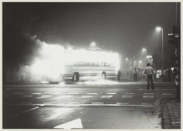 Schwarz-weiß-Foto eines brennenden Busses am Straßenrand mit einer Gruppe von Menschen auf dem Gehweg rechts und Straßenlaternen im Hintergrund.
