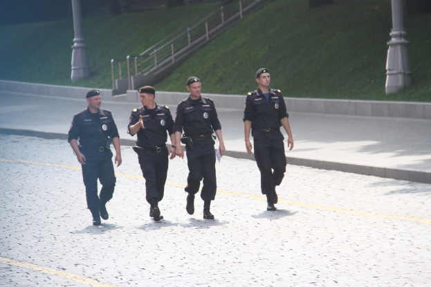 Drei Polizisten in Uniform und Mützen, die eine Straße mit Pfählen, Geländern, Treppen und Gras im Hintergrund entlanggehen.