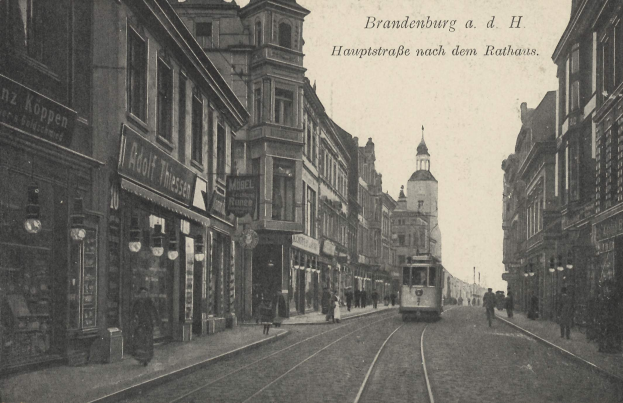 Ein altes Schwarz-Weiß-Foto einer Stadtstraße in Brandenburg, Deutschland, mit einer Straßenbahn auf den Schienen, Menschen auf dem Gehweg, Gebäuden mit Fenstern, Schildern und Straßenlaternen unter einem bewölkten Himmel.