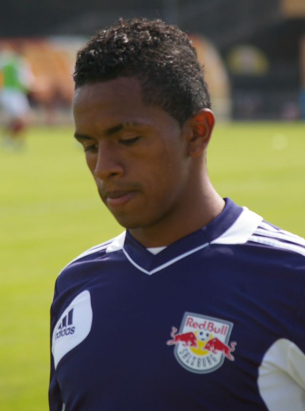 Junger Mann in blauer T-Shirt auf einem Fussballfeld, der wie ein Flügelspieler mit kurzen, dunklen Haaren und entschlossener Miene aussieht.