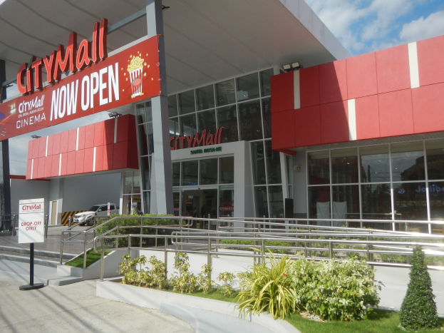 Stadteingangsschild des City Malls mit der Aufschrift "City Mall jetzt geöffnet" mit umliegender Vegetation, Menschen, einem Auto und einem bewölkten Himmel.