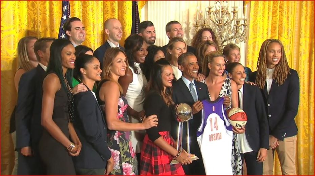 Präsident Obama und First Lady Michelle Obama posieren mit dem Damen-Basketball-Team im Oval Office des Weißen Hauses, halten einen Basketball, eine Trophäe und lächeln in die Nähe einer Flagge, Vorhänge und einer Kerzenleuchter.