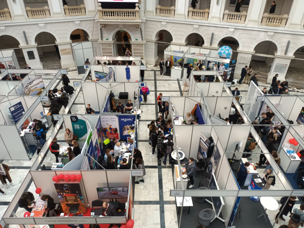 Große Besuchermenge auf einer internationalen Fachmesse mit Ständen, Bannern, Tischen, Stühlen und Luftballons in einem Raum mit architektonischen Bögen und Säulen.